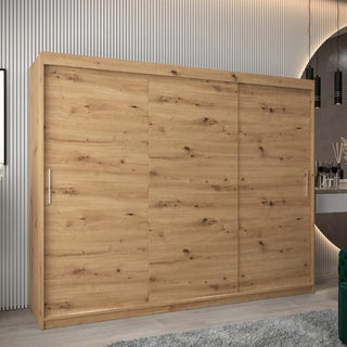 The Tavira Wooden Wardrobe 3 Sliding Doors 250cm in Artisan Oak stands against a vertically paneled wall, featuring metal handles. A soft rug is on the floor, with a mirror and decorative items visible to the right.