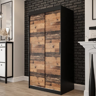 The Topeka Wooden Wardrobe with 2 Sliding Doors in Dark Oak features a black frame and rustic panels, standing against a black brick wall on light wood flooring for stylish bedroom storage. A white dresser and fireplace are partially visible nearby.