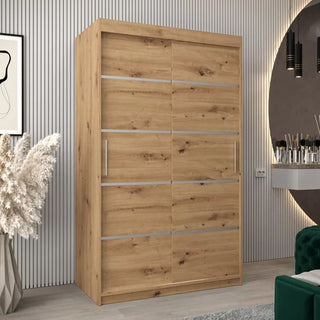 The Vevey I Wooden Wardrobe with 2 sliding doors (120cm, Artisan Oak) features metallic handles and is showcased in a modern room with ribbed wall panels, a round mirror, wall-mounted vanity, green chair, and dried grass in a vase.