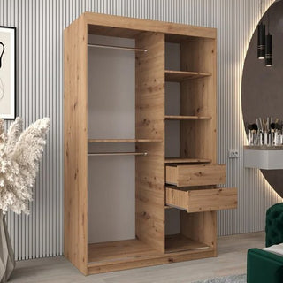 The Vevey I Wooden Wardrobe with 2 sliding doors (120cm, Artisan Oak) is showcased in a modern room with a large round mirror, green chair, pampas grass arrangement, and striped wall paneling.