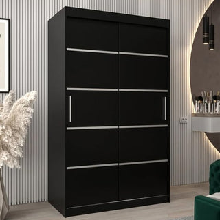 The Vevey I Wooden Wardrobe with 2 sliding doors in black (120cm) features horizontal silver handles and stands against a vertically-striped wall in a modern room, accompanied by a decorative plant and vanity table.