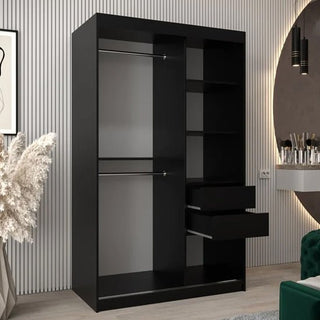 The Vevey I Wooden Wardrobe with 2 sliding doors (120cm, black) features two hanging rods, shelves, and two drawers, standing in a stylish room with light wood floors, decorative plants, and a round mirror above a vanity.