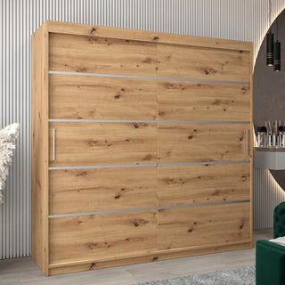 The Vevey I Wooden Wardrobe, 200cm in artisan oak, stands against a grooved wall. It features 2 sliding doors with a silver handle on the left and horizontal metal accents, all in a natural finish.