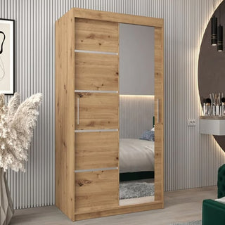 The Vevey II Mirrored Wardrobe 2 Sliding Doors 100cm in Artisan Oak stands in a modern bedroom with a striped wall, decorative pampas grass, and a corner vanity.