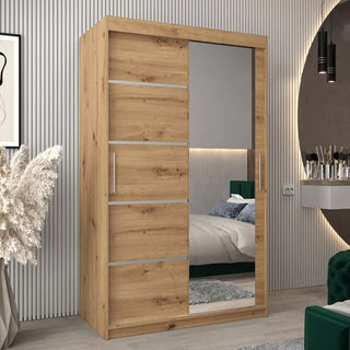 The Vevey II Mirrored Wardrobe 2 Sliding Doors 120cm in Artisan Oak features one mirrored and one wood panel door, elevating the style of a modern bedroom with light gray walls, a bed, and chic decor.
