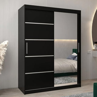 The Vevey II Mirrored Wardrobe, 150cm in black with two sliding doors—one mirrored and one with silver accents—stands in a modern bedroom with a striped wall, round mirror, and bed in the background.
