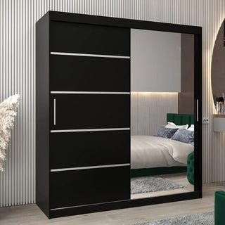 The Vevey II Mirrored Wardrobe with 2 sliding doors (180cm, black) features silver accents and a large mirrored door, reflecting the bed, rug, and green upholstered headboard in a stylish contemporary bedroom.