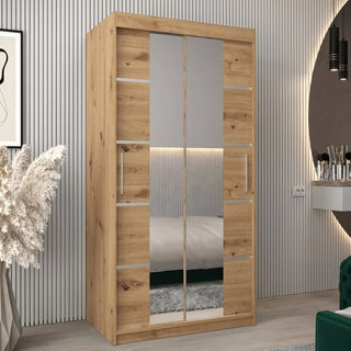 The Vevey III Mirrored Wardrobe with 2 sliding doors in artisan oak (100cm) is showcased in a modern room with vertical wall paneling, a plant in a vase, and a round mirror above a small vanity.
