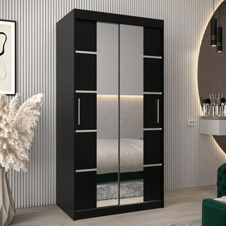 The Vevey III Mirrored Wardrobe 2 Sliding Doors 100cm in Black features mirrored panels and silver handles, stands against a grooved wall, and is placed beside a vase with pampas grass, a floating desk with cosmetics, and a circular wall light.