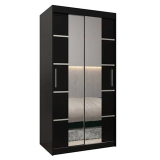 The Vevey III Mirrored Wardrobe features 2 black sliding doors, mirrored panels, and rectangular metal handles with geometric accents. The mirrors reflect a bed, wall, and rug in a modern bedroom decor. Width: 100 cm.