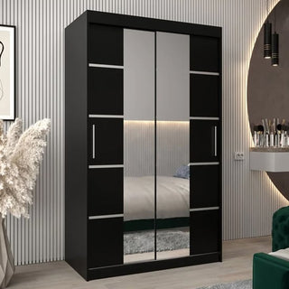 The Vevey III Mirrored Wardrobe with 2 sliding doors (120cm, black) stands in the bedroom. Vertical handles and horizontal accents add style. Its mirrored doors reflect a bed, rug, wall mirror, pampas grass, and green chair.