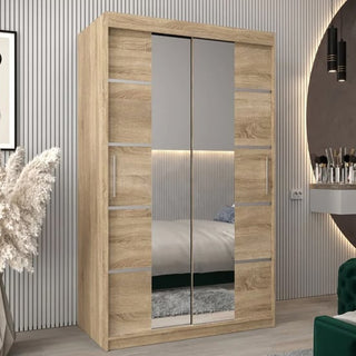 The Vevey III Mirrored Wardrobe with 2 sliding doors in Sonoma Oak (120cm) stands against a light, vertically-striped wall. It features metal handles, and its mirror reflects part of the bed and rug in the room.