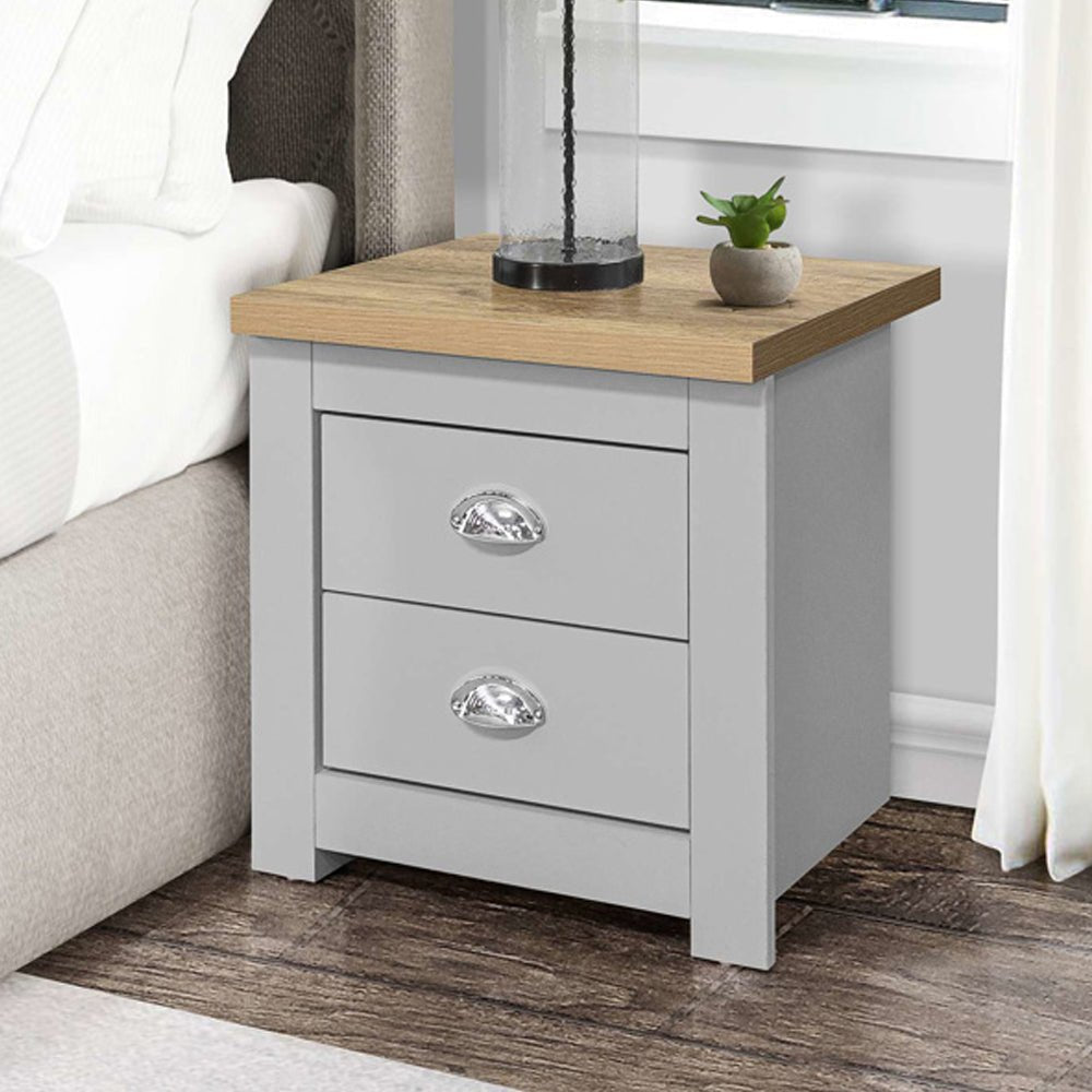 Grey wooden bedside cabinet with 2 drawers and silver handles, oak-effect top, next to a bed.