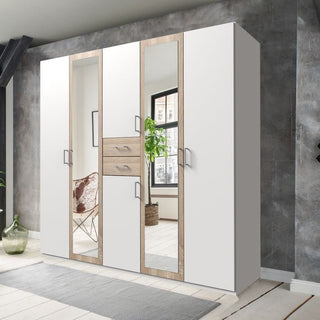 The DAKAR 5 Door Wardrobe, 225cm in white and oak, features mirrored doors, four white panels, two central wooden drawers, and silver handles, complementing a room with a gray accent wall and wooden flooring.