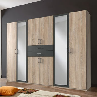The Wardrobe - DAKAR 6 Door Wardrobe Oak and Graphite 270cm stands against a dark wall, featuring mirrored doors, three central drawers, six metal-handled doors, and a wide design. A rug and cushions are placed in front.