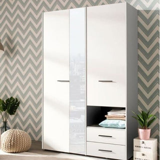 The HIGHAM 3 Door Mirrored Wardrobe in white (127.5cm) stands against a zigzag-patterned wall, featuring spacious storage. A round woven pouf, green plants, and light wood flooring enhance the room’s charm.