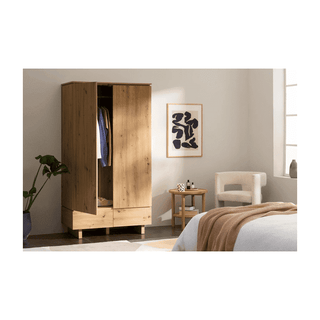 The TUNA 2 Door Wardrobe in Artisan Oak Effect (99cm) stands with partially open doors next to a side table, white armchair, and beige-bedded bed in a bright, minimal bedroom. A framed abstract artwork adorns the wall.